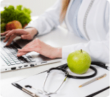 Nutricionista digita em um laptop. Em primeiro plano, uma maçã verde repousa sobre um estetoscópio e uma prancheta