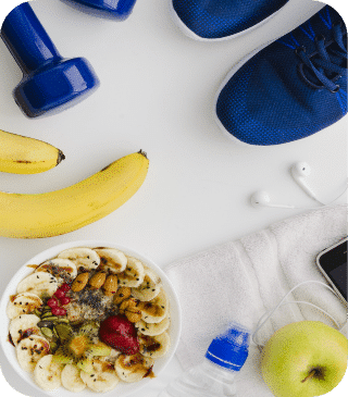 Layout com tema fitness, apresentando halteres azuis, uma tigela de smoothie saudável coberta com frutas e nozes, bananas, tênis azuis, fones de ouvido, uma toalha, smartphone, maçã verde e garrafa de água em uma superfície branca