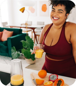Uma mulher sorri, segurando um copo de suco de laranja, em frente a um liquidificador e frutas fatiadas, incluindo laranjas, pitaya e mamão