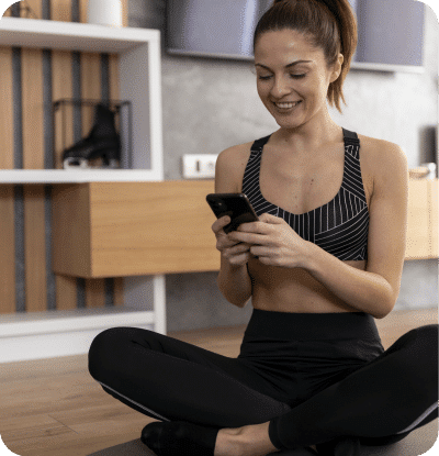 A mulher em uma roupa de treino sentada de pernas cruzadas no chão na sala da sua casa, sorrindo para o telefone