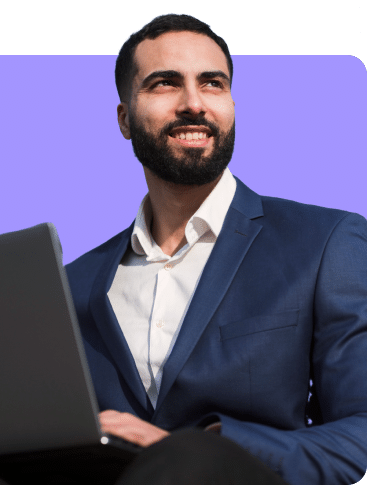 Um homem com barba, de terno azul e camisa branca, sorri enquanto segura um laptop. O fundo é de uma cor roxa sólida.