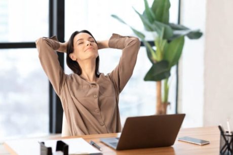 Uma mulher de blusa bege se recosta na cadeira com as mãos atrás da cabeça, personificando a essência da saúde mental no trabalho.