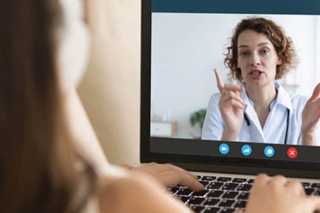 Uma pessoa em videochamada exibida na tela de um laptop, com outra pessoa assistindo de trás, ilustrando a telemedicina para empresas