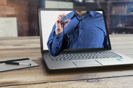 Um laptop sobre uma mesa de madeira exibe a imagem de uma pessoa de camisa azul segurando um estetoscópio ilustrando a telessaúde no Brasil